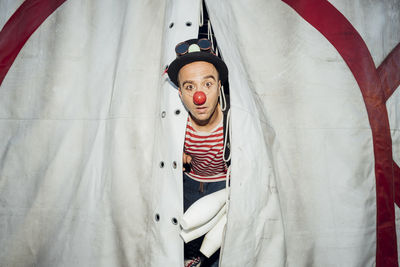 Male clown with hat and juggling pin peeking through circus tent entrance