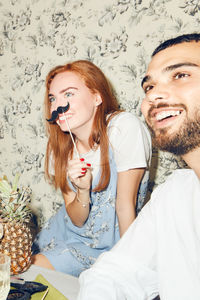 Smiling redhead woman holding mustache prop while sitting by male friend during party at home