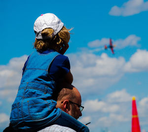 Rear view of daughter sitting on father shoulders against sky