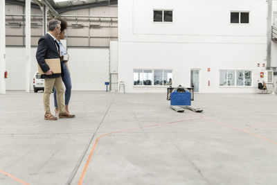 Businessman holding laptop standing with businesswoman looking at pallet jack in factory