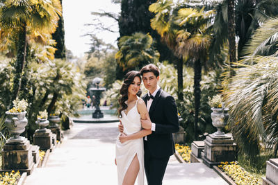 Happy lovers the bride and groom in wedding outfits walk among plants and palm trees in the old park