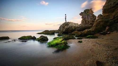 Man standing on rock at shore against sky during sunset