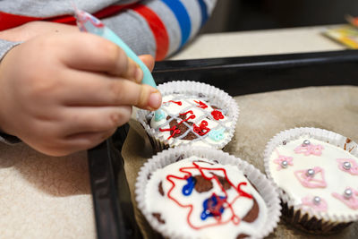 A child squeezes colored frosting from a tube onto chocolate brown cupcakes covered  white frosting.