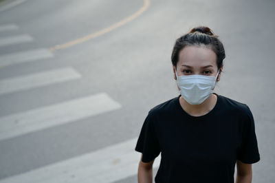 Front view young woman wear protection mask in black casual wear standing near pedestrian crossing