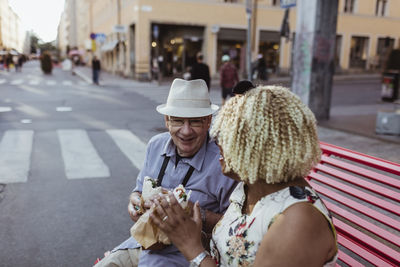 Senior couple having food while sitting on bench in city