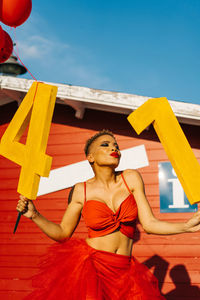 Trendy black female with decorative numbers and red balloons standing on walkway against construction during birthday party in sunlight