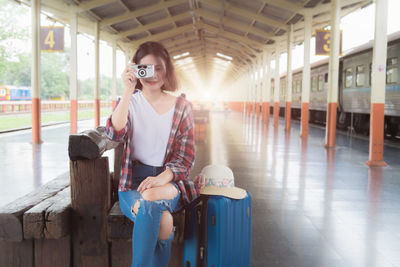 Portrait of young woman photographing while sitting on bench at railroad station