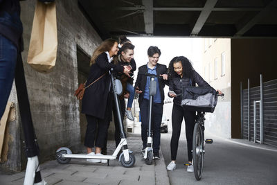 Happy male and female teenage friends with electric push scooters looking at mobile phone below bridge