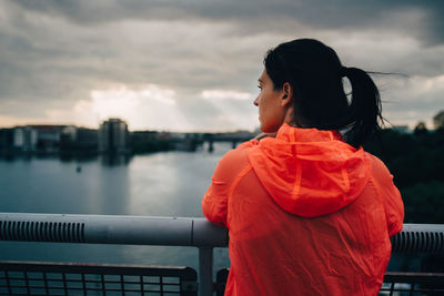 Rear view of sportswoman in raincoat looking at city while standing on footbridge over sea during sunset