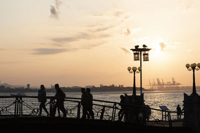 Silhouette people on street by sea against sky during sunset