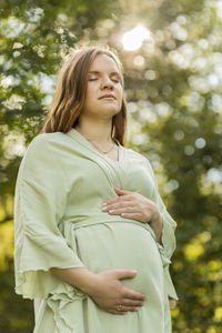 Young pregnant female holds hands on big belly, closed eyes, green trees,sky, sunlight on background