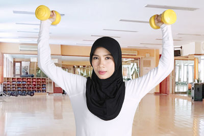 Portrait of young woman lifting dumbbells while standing in gym