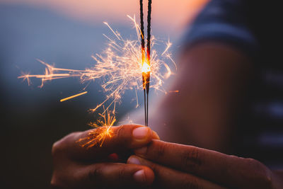 Cropped hand holding lit sparkler during sunset