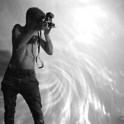 Shirtless man photographing with instant camera against sunlight on wall