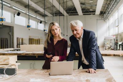 Businesswoman discussing over laptop with businessman leaning on workbench in factory