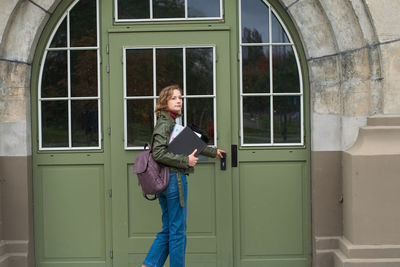 Female student with a backpack holds notes and opens the door to the college