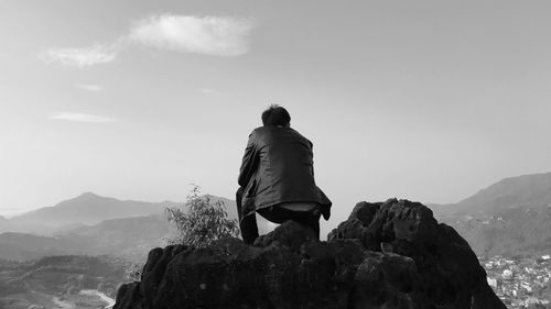 Rear view of man crouching on rock against sky