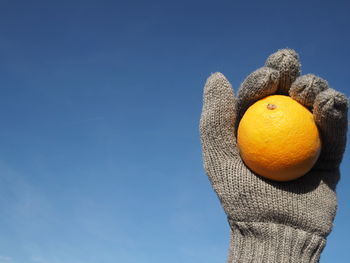 Cropped image of hand wearing gloves holding orange against sky