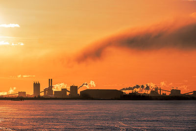 Factory by sea against orange sky