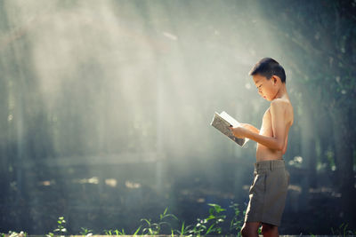 Shirtless boy reading book while standing outdoors