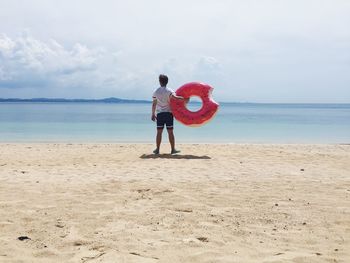 Full length rear view of man with donut shape life belt standing at beach against sky