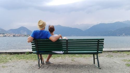 Rear view of woman sitting on bench by lake against sky