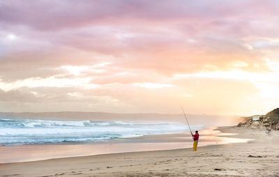 Man with fishing rod standing on beach against sky during sunset