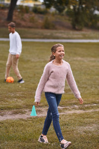 Smiling sister walking on grassy field while brother playing soccer in background at park