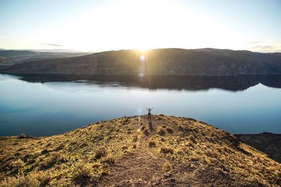 Distant view of person standing on cliff by lake and mountain against sky during sunset