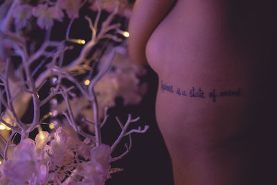 Midsection of shirtless woman with tattoo by illuminated flowers