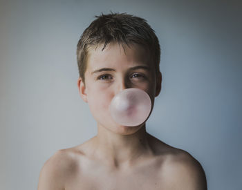 Portrait of shirtless playful boy blowing bubble gum against wall at home