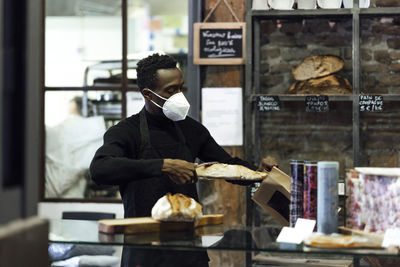 Male bakery owner packing bread in paper bag while standing at bakery