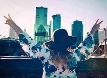 Rear view of woman with arms raised showing peace sign while standing in city