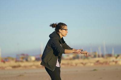 Side view of young woman gesturing while standing at beach against clear blue sky