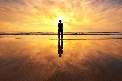 Rear view of man standing on shore at beach against sky during sunrise