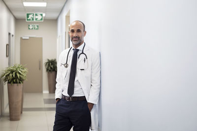 Portrait of smiling doctor with hands in pockets leaning on wall at hospital