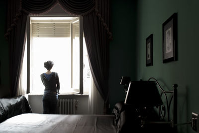 Rear view of woman standing near window in bedroom