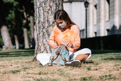 Young female student in mask sitting under tree outside university and rummaging through backpack on sunny day in madrid, spain