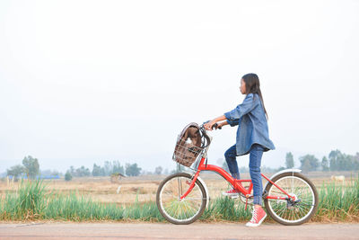 Side view of teenage girl with bicycle on roadside by field against clear sky