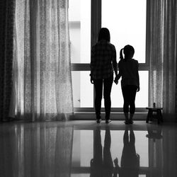 Rear view of siblings standing on floor by window at home