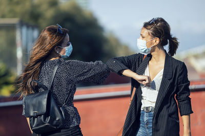 Friends wearing face mask giving elbow bumps while standing in city