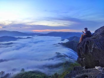 Side view of man looking at cloudscape while sitting on mountain during sunset