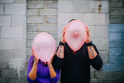Close-up of man and woman holding balloons over face while standing against wall