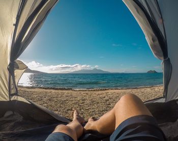 Low section of person relaxing on sea shore against sky