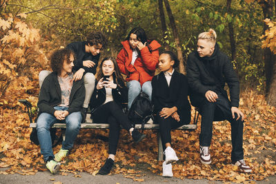 Multi-ethnic friends looking at teenage girl using mobile phone while sitting on bench at park