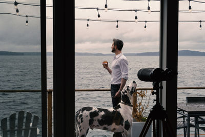 Man holding drink in glass with dog standing by railing and sea against sky