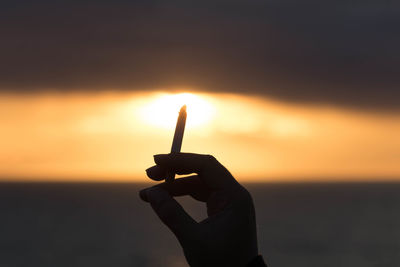 Cropped hand holding cigarette against sea during sunset