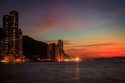 Illuminated buildings by sea against sky at sunset