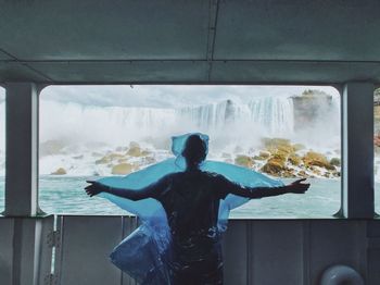 Boy in plastic standing by window against waterfall