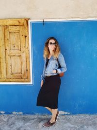 Full length of mid adult woman wearing sunglasses standing against blue wall